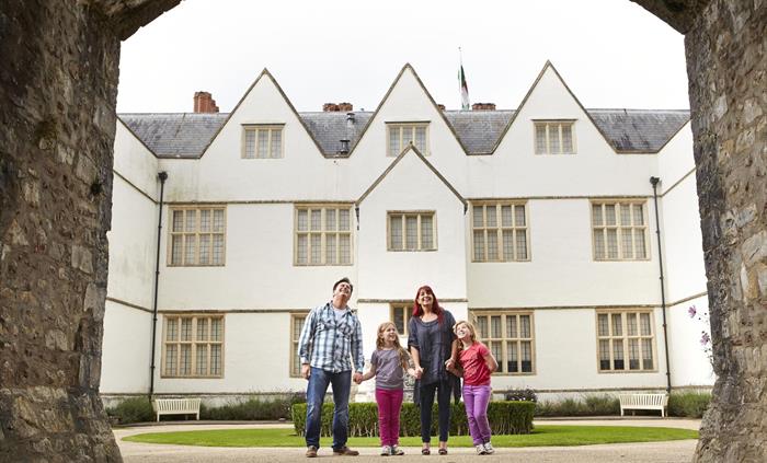 a family of two parents and two young girls stand beneath a high stone archway. in the background is a large white building, with lots of leaded windo